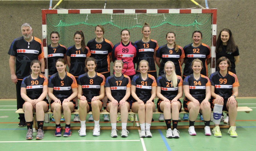 Frauen-Handballmannschaft posiert in Sporthalle, Spielerinnen in schwarzen Trikots mit Nummern, Trainer links.