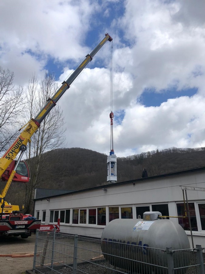 Kran hebt Klimaanlage auf Gebäude in ländlicher Umgebung, umgeben von Bäumen und Bergen, unter bewölktem Himmel.