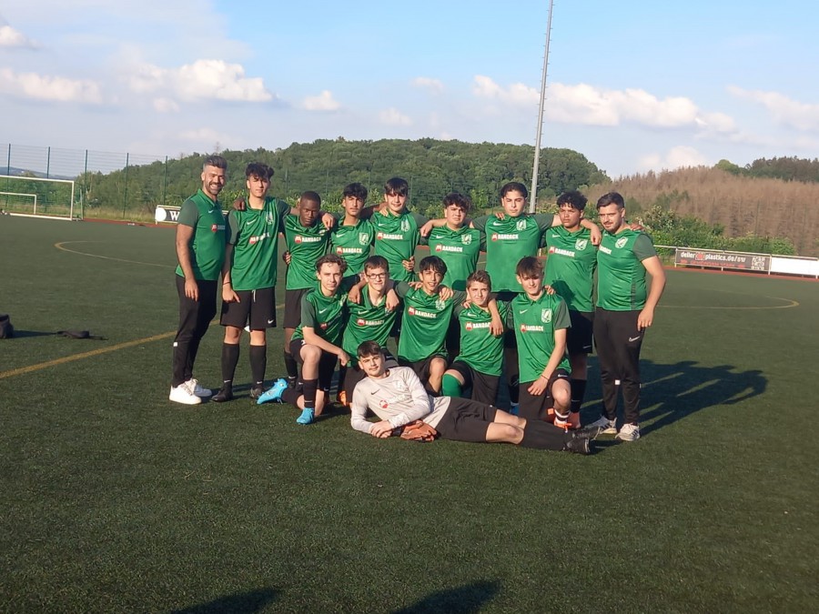Fußballmannschaft in grünen Trikots posiert auf dem Platz an einem sonnigen Tag, mit Bäumen im Hintergrund.