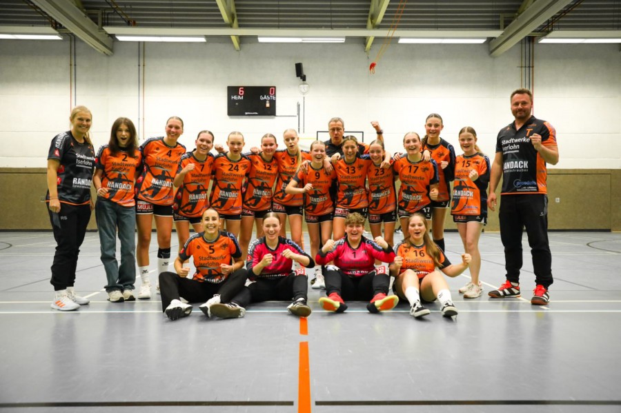 Frauen-Handballteam in orangenen Trikots jubelt in der Turnhalle nach einem Spiel oder Training, Gruppenfoto mit Trainer.