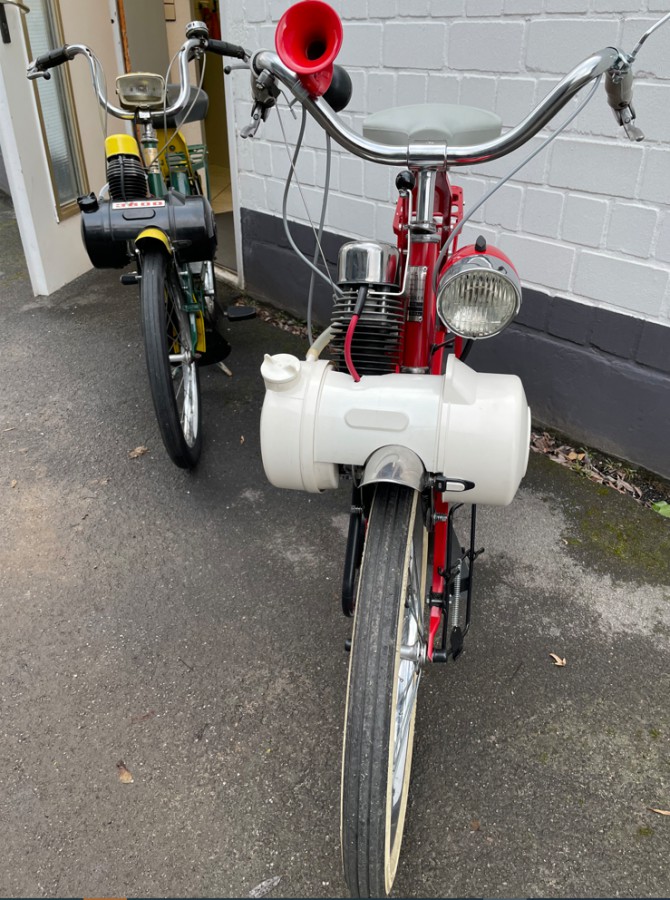 Velosolex-Restaurierungsprojekt_Doerken Zwei Vintage-Mofas vor einer weißen Ziegelwand, eins rot mit Hupe, das andere schwarz-gelb, seitlich vor einer Tür geparkt.